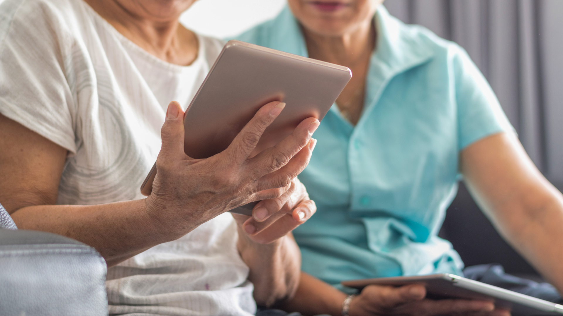 Two older women seated on a couch, engaged in viewing content on a tablet together.