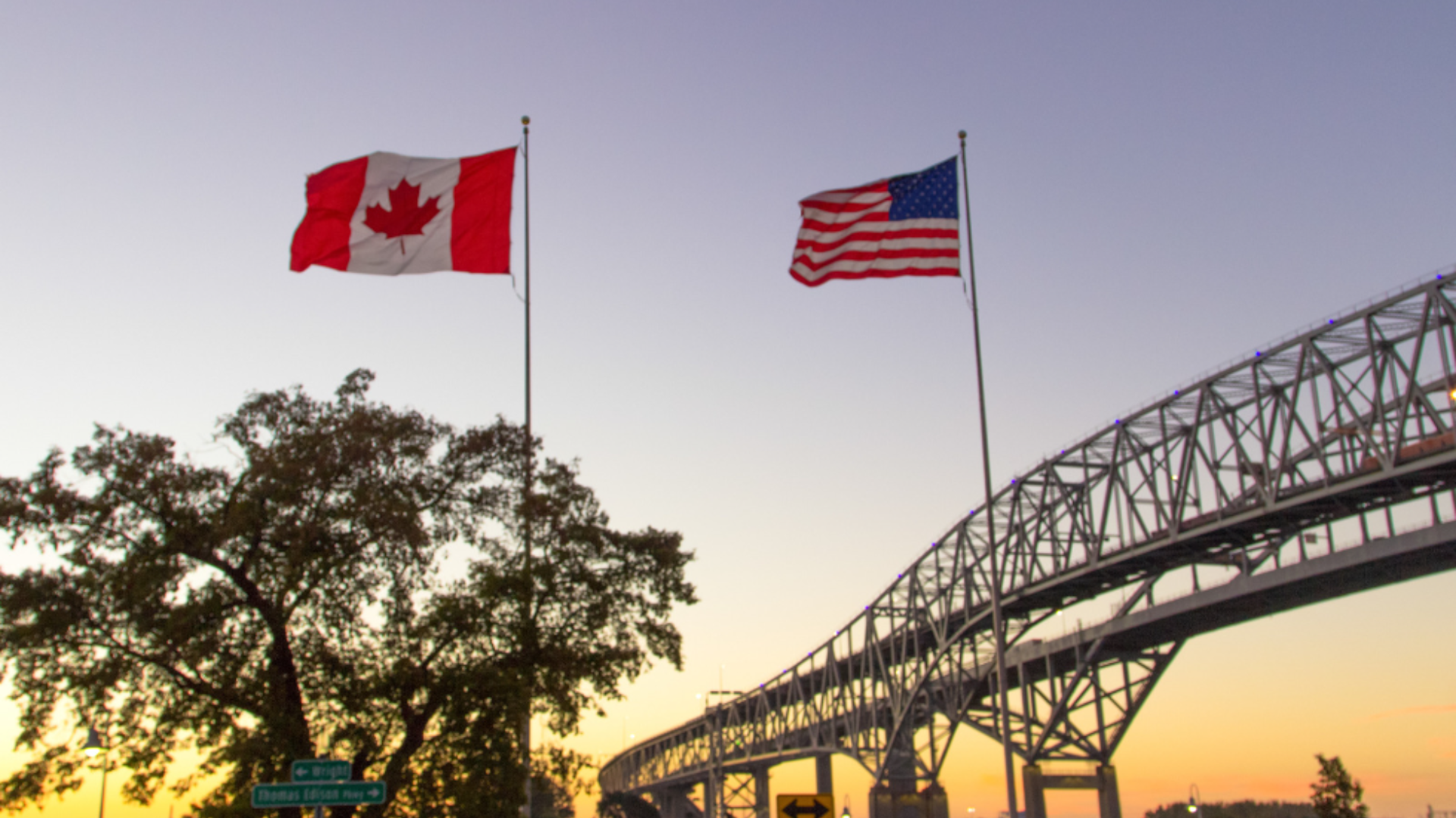 A Canadian flag and a United States flag flutter in front of a bridge.