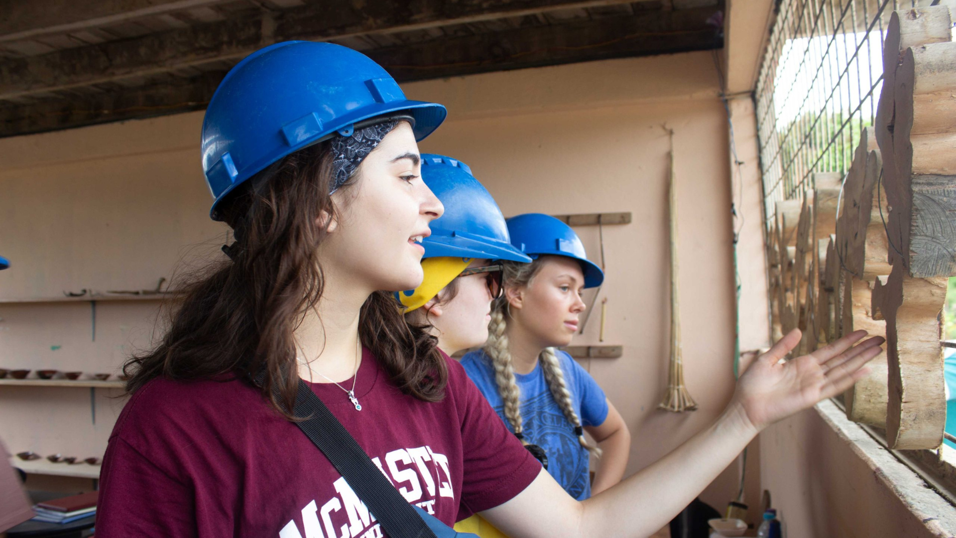 Three girls wearing hard hats examine a wall, indicating a hands-on educational experience.