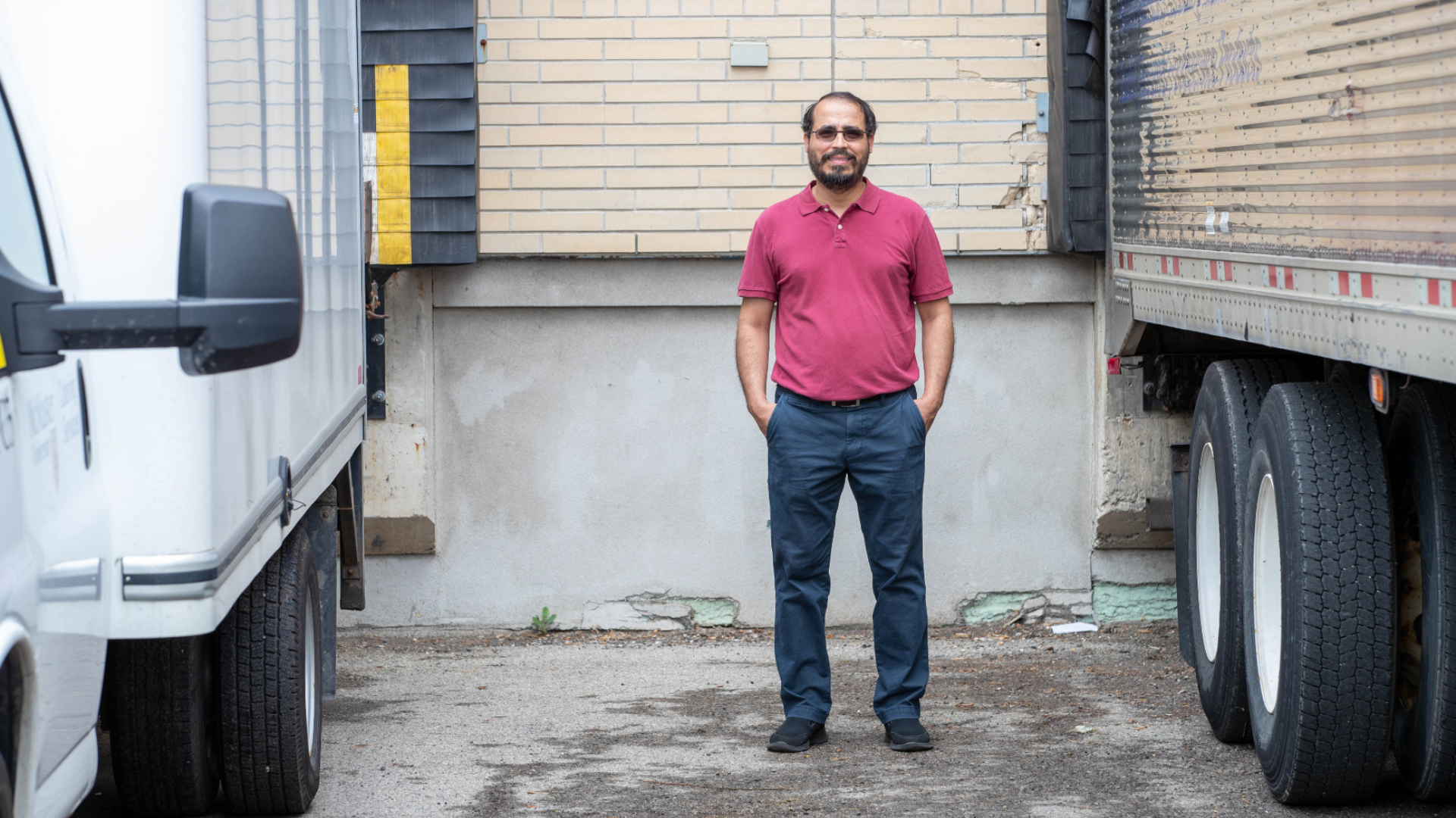 Elkafi Hassini stands confidently between two delivery trucks, highlighting his involvement in the transportation supply chain.