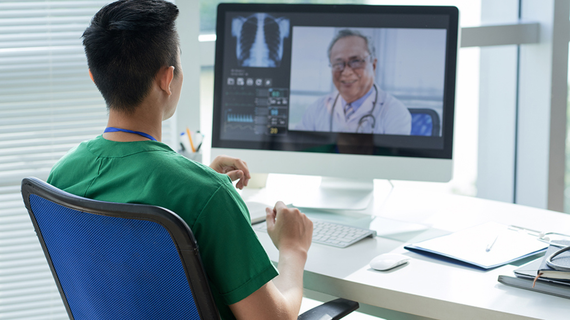 A man wearing a green shirt is seated at a desk, interacting with a computer screen for a virtual doctor consultation.