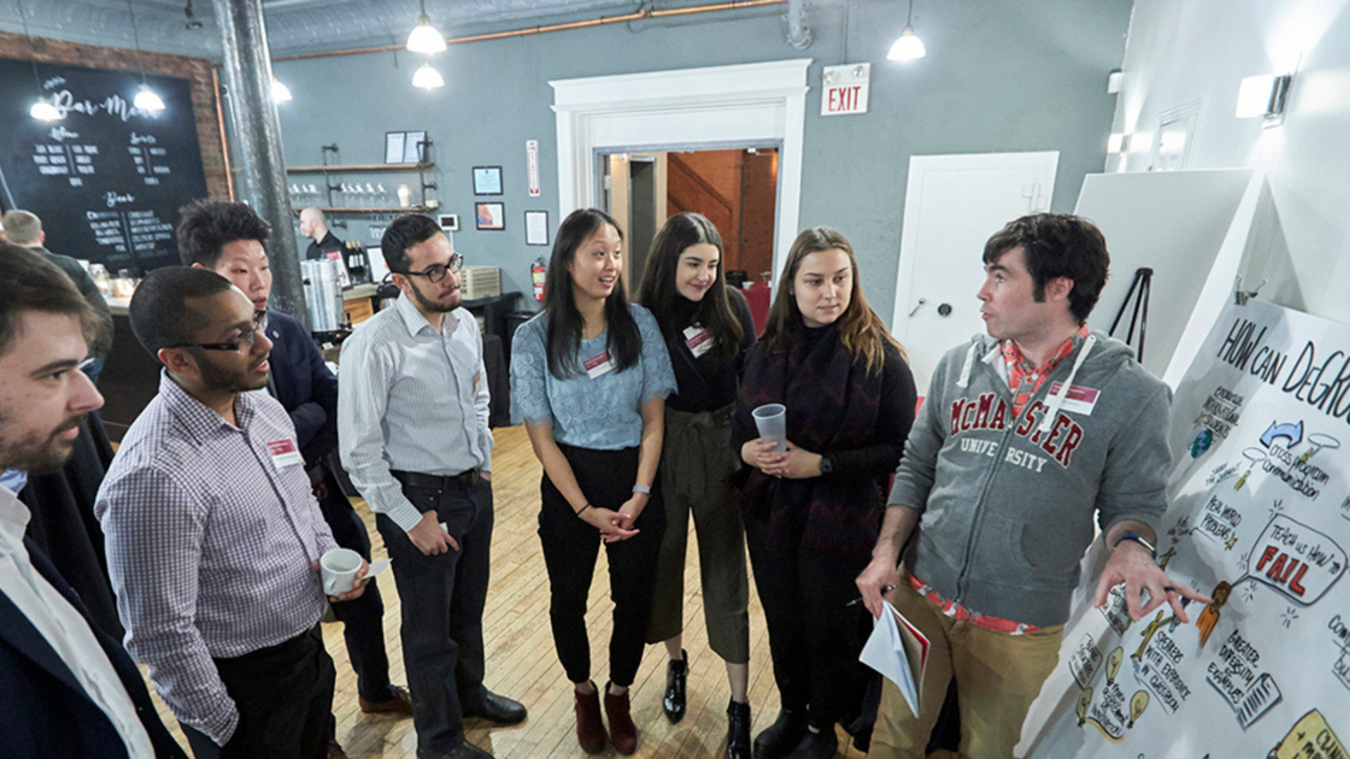 A DeGroote staff member engages with potential students near a poster board at an open house event.