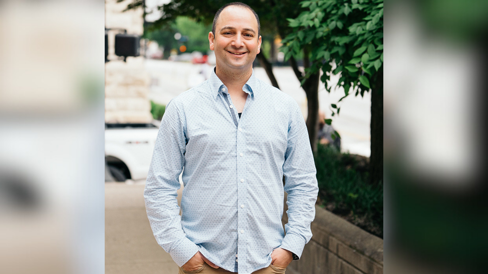 Tal Dehtiar, dressed in a blue shirt and tan pants, smiles while standing on a sidewalk.