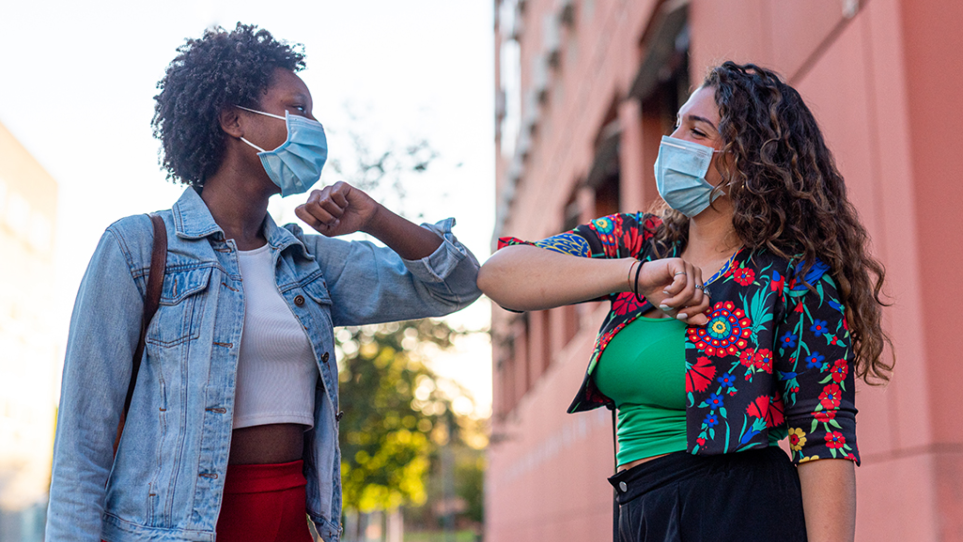 Two women wearing face masks greet each other with an elbow handshake while standing outdoors.