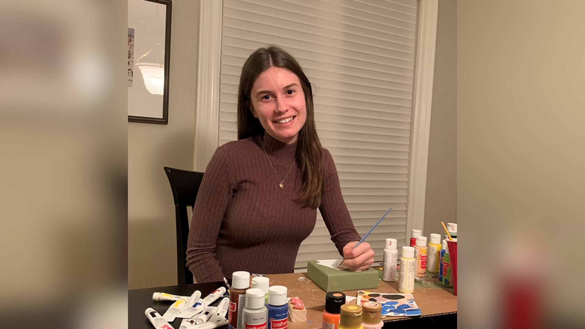 Rachel West, a woman at a table, smiles while holding a paintbrush next to a box of paints and various art supplies.