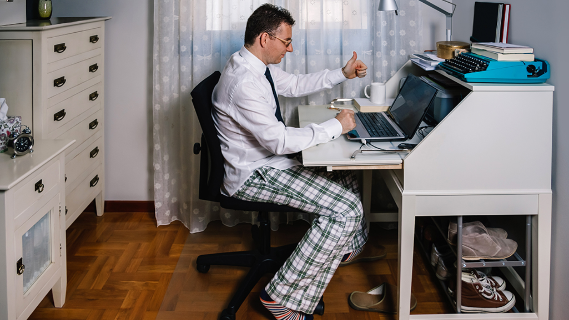 A man in pajama pants and a white dress shirt works at a desk with a laptop, smiling and giving a thumbs up.