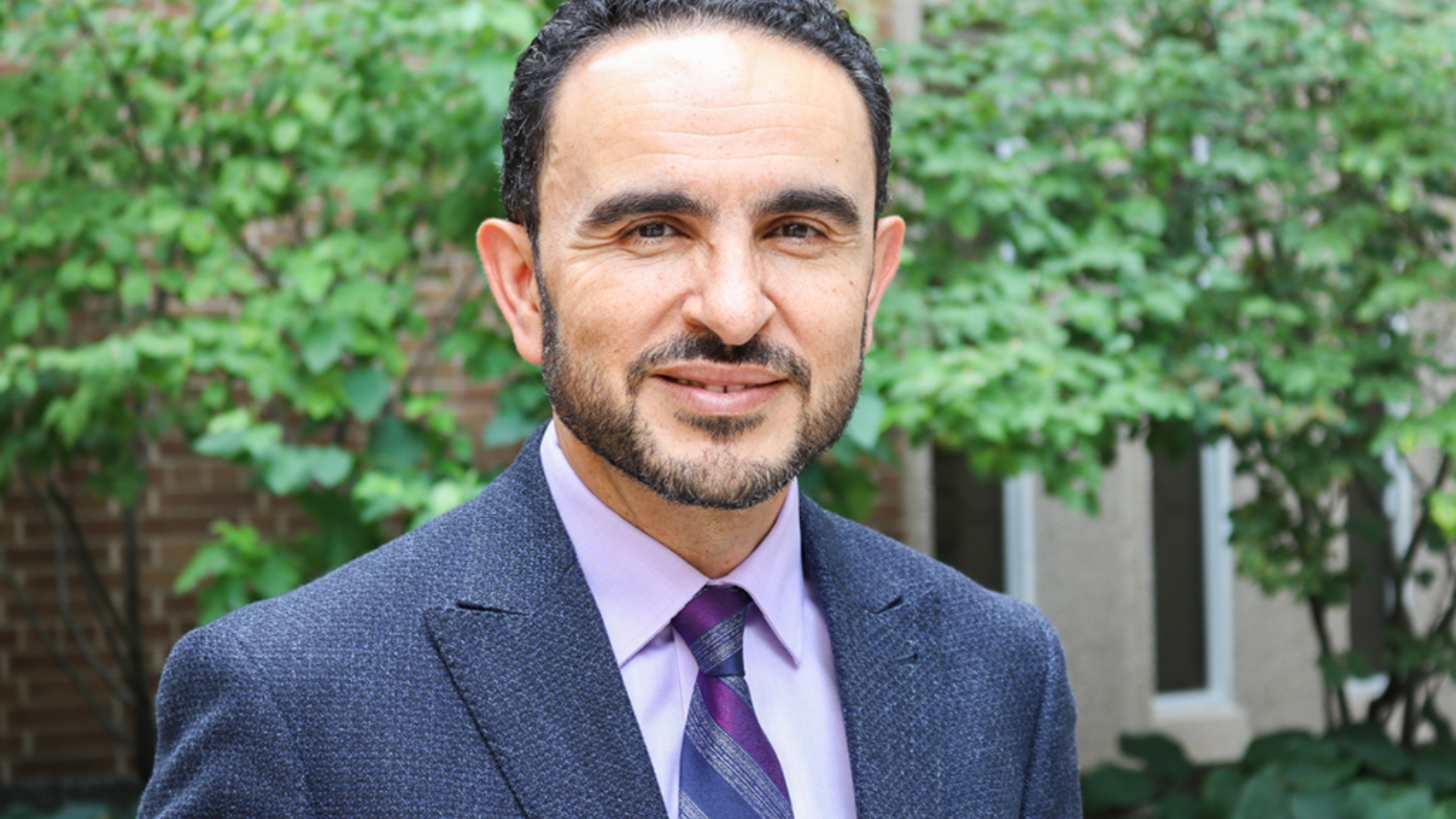 Khaled Hassanein, dressed in a suit and tie, smiles while standing amidst a backdrop of vibrant trees.