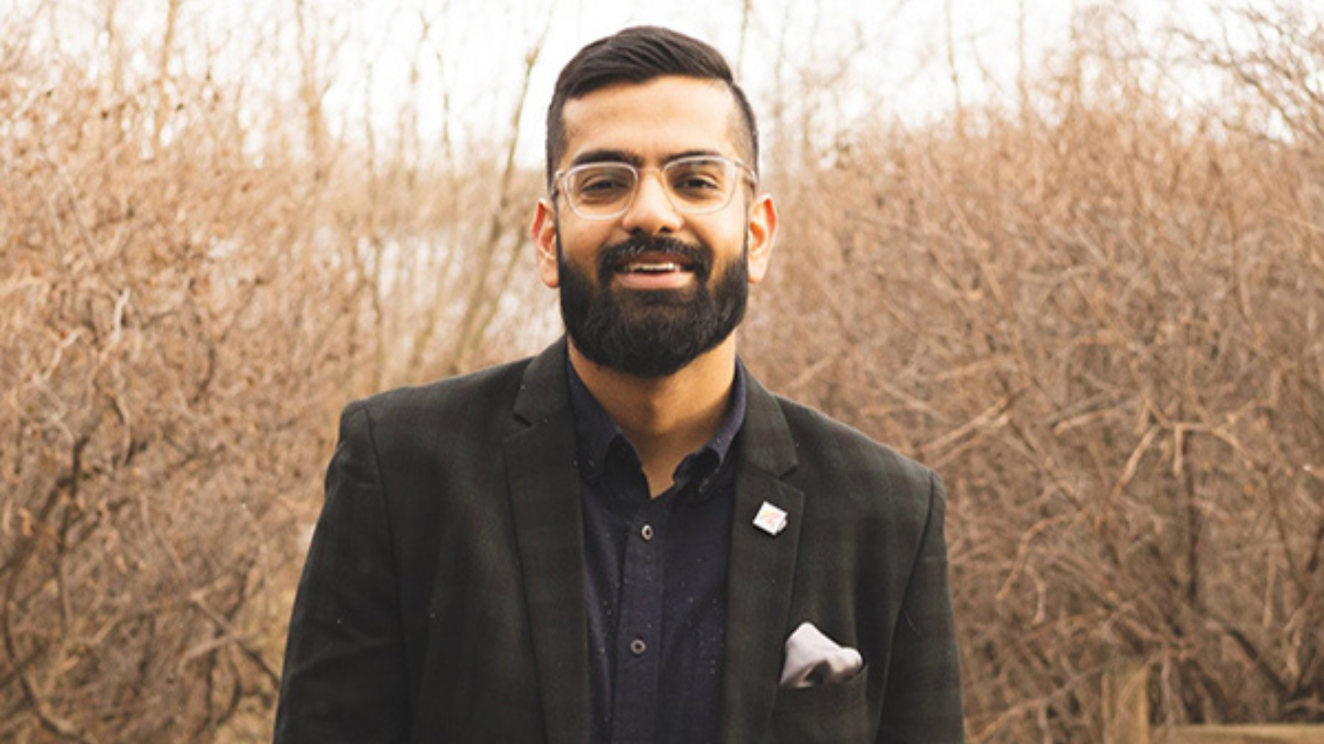 A smiling man with a beard and glasses stands in front of lush green trees, exuding a warm and friendly demeanor.