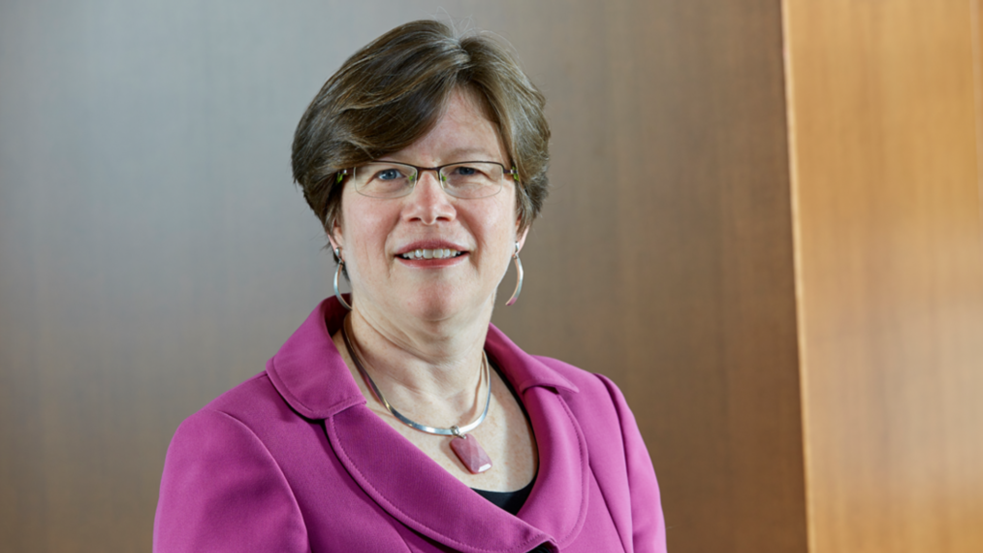 Gillian Mulvale, wearing glasses and a pink jacket, smiles while standing against a wooden wall.