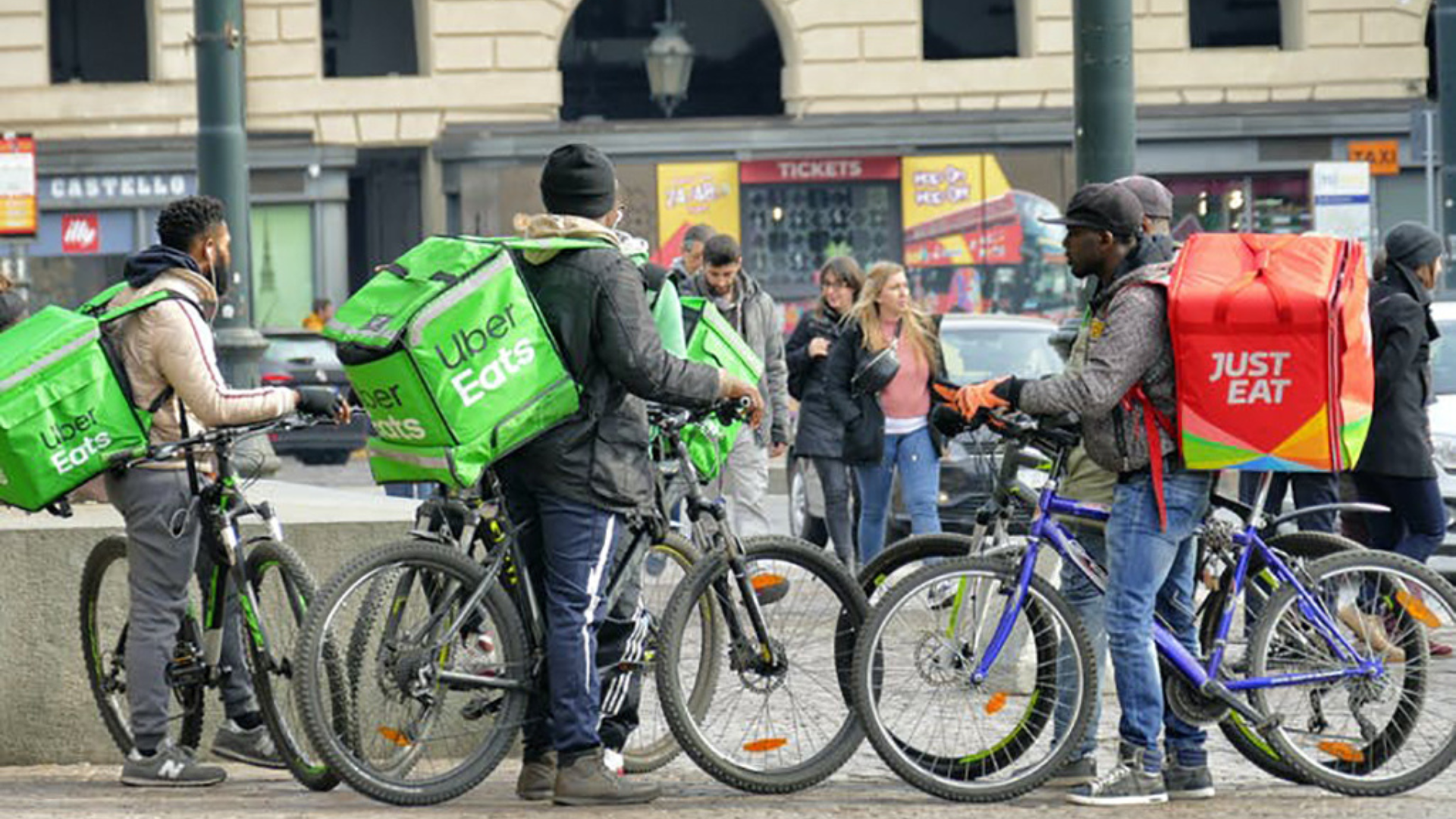 Five Uber Eats gig workers with bags on their bikes stand in front of a building, ready for their next delivery.