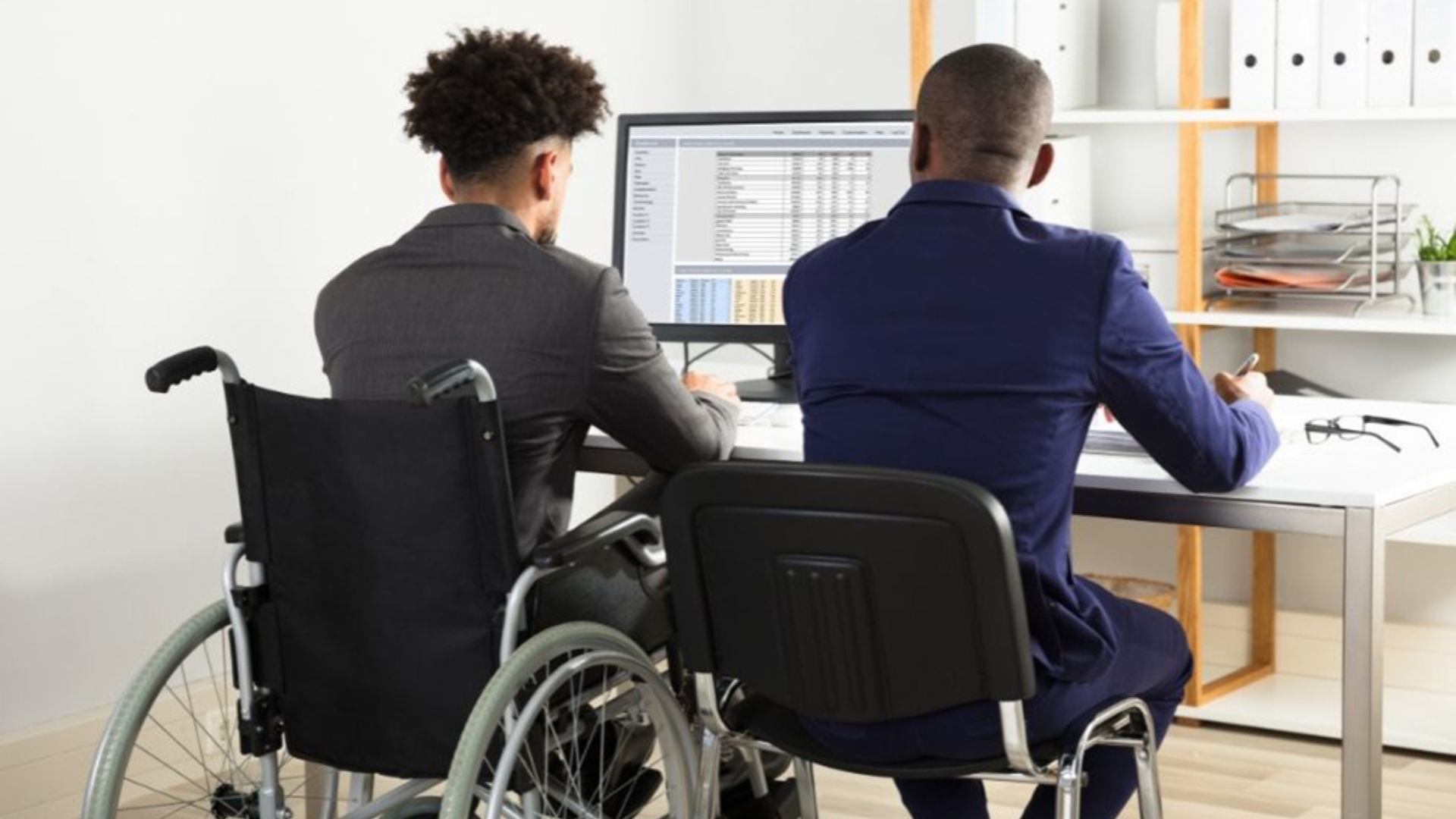 Two men, one in a wheelchair and one in a regular chair, collaborate at a desk in a professional office environment.