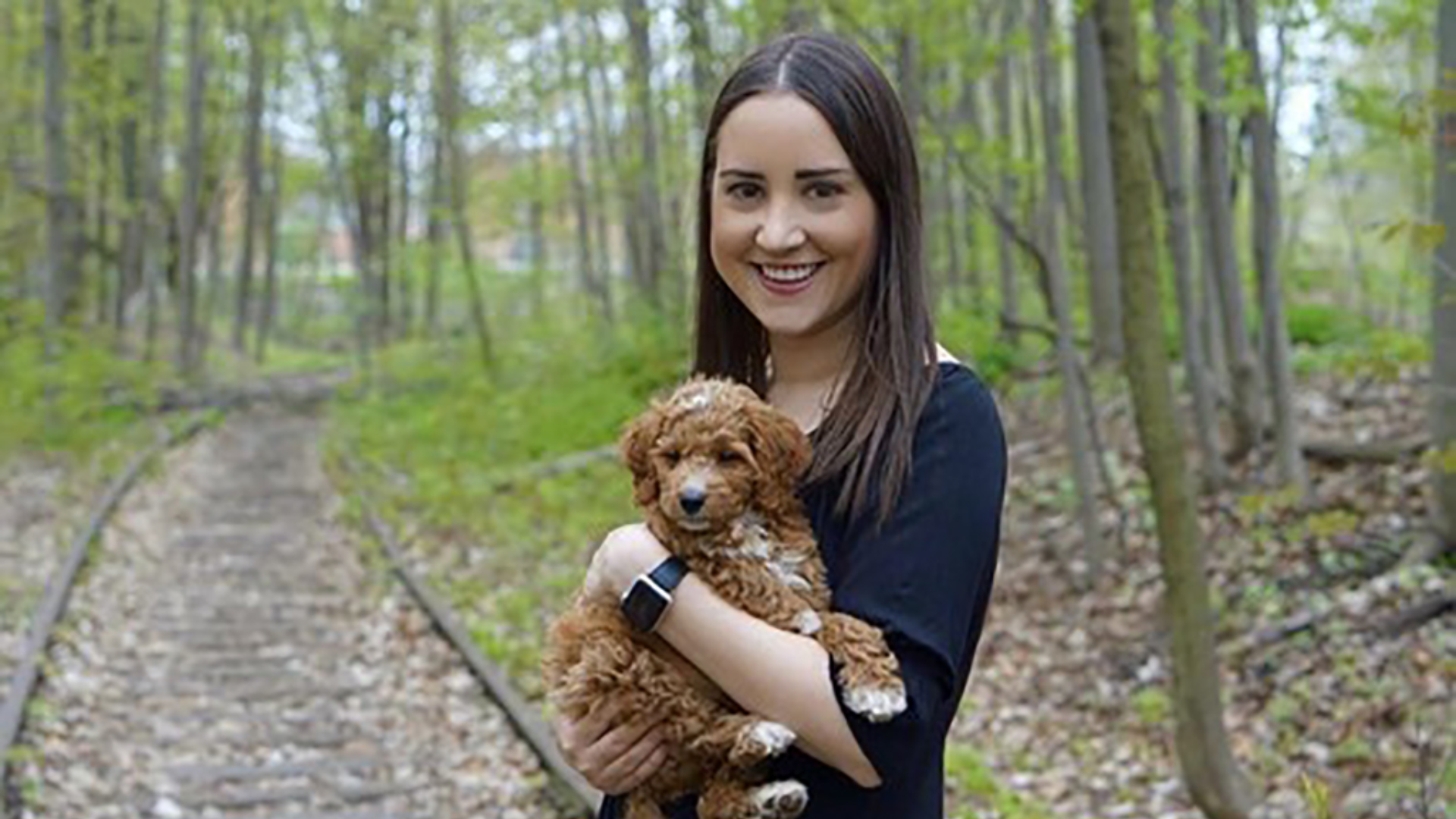 Christine Peral smiles while holding her dog on a scenic hiking trail.