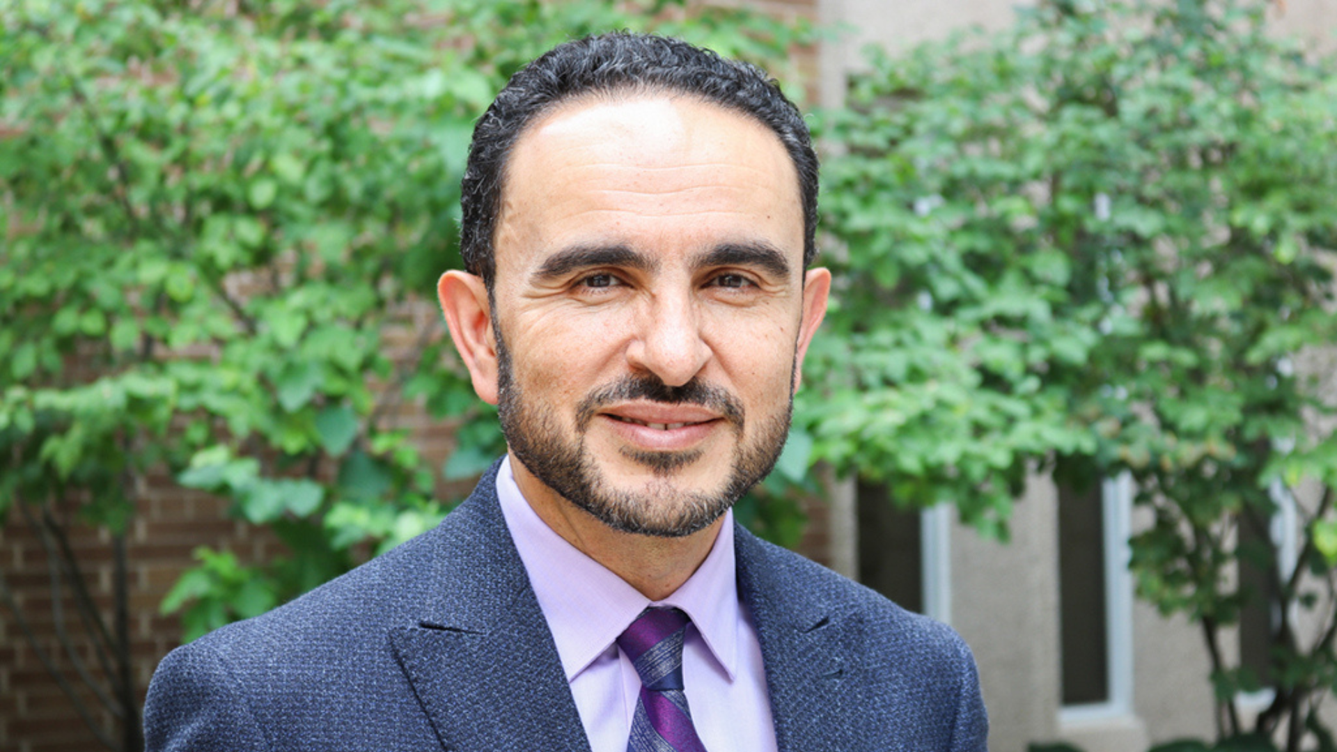 Dean Khaled Hassanein, smiling, stands in front of trees, dressed in a suit and tie.