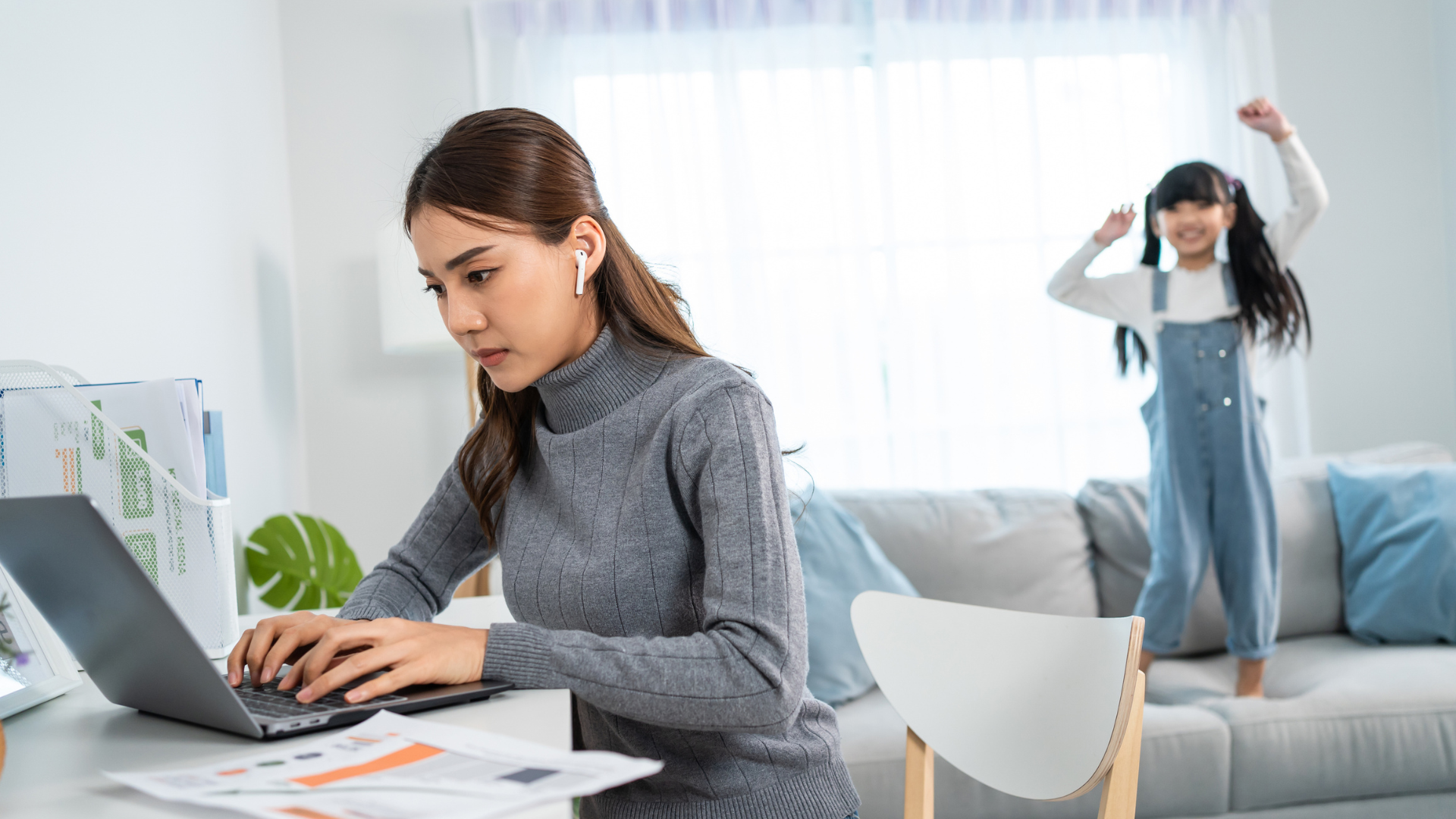 A woman working at home at her desk with a laptop while a small child jumps on the couch in the background.