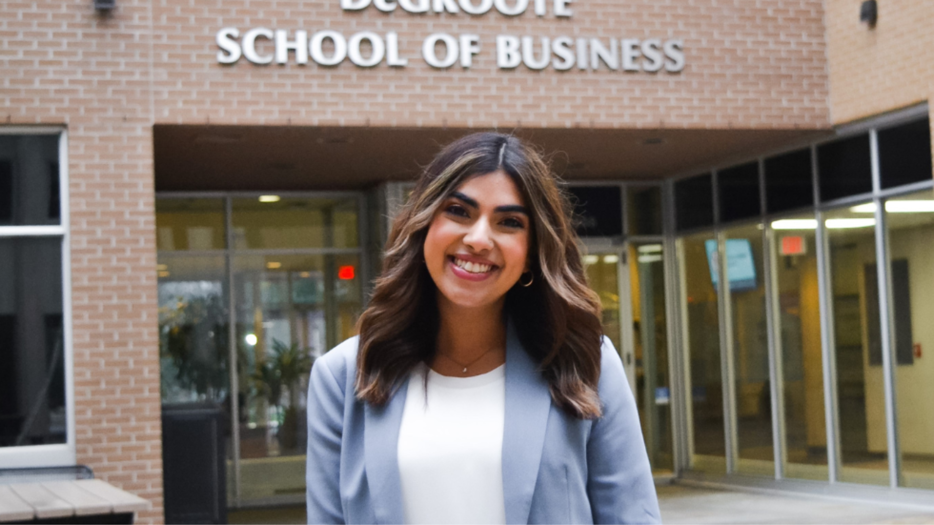 Momina Qureshi smiles in a white shirt and light blue jacket, standing in front of the DeGroote School of Business building.