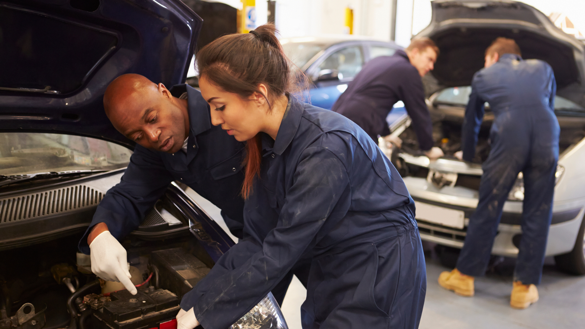 Two individuals collaborating on a car repair inside a well-equipped garage, surrounded by tools and automotive parts.