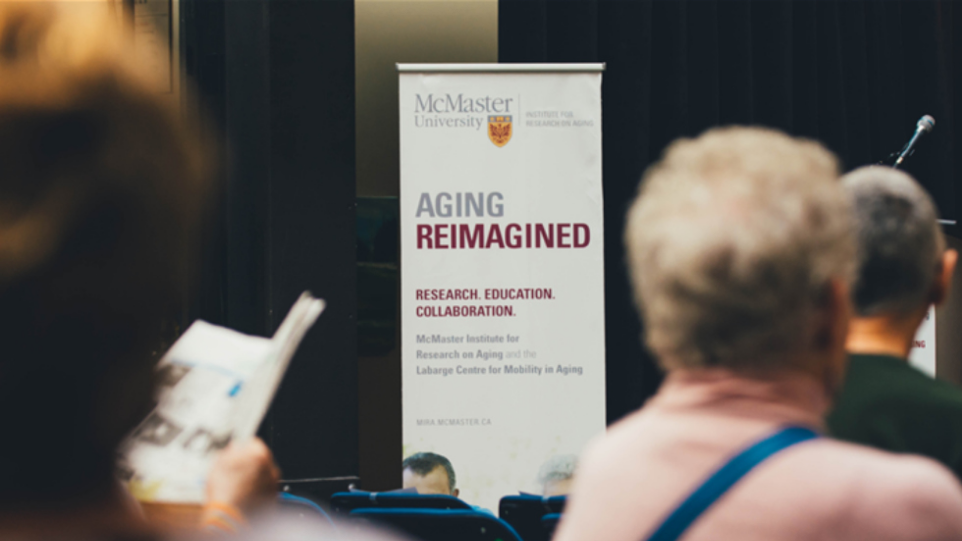 Sign for McMaster University’s Aging Reimagined event in focus, with attendees seated out-of-focus around it.