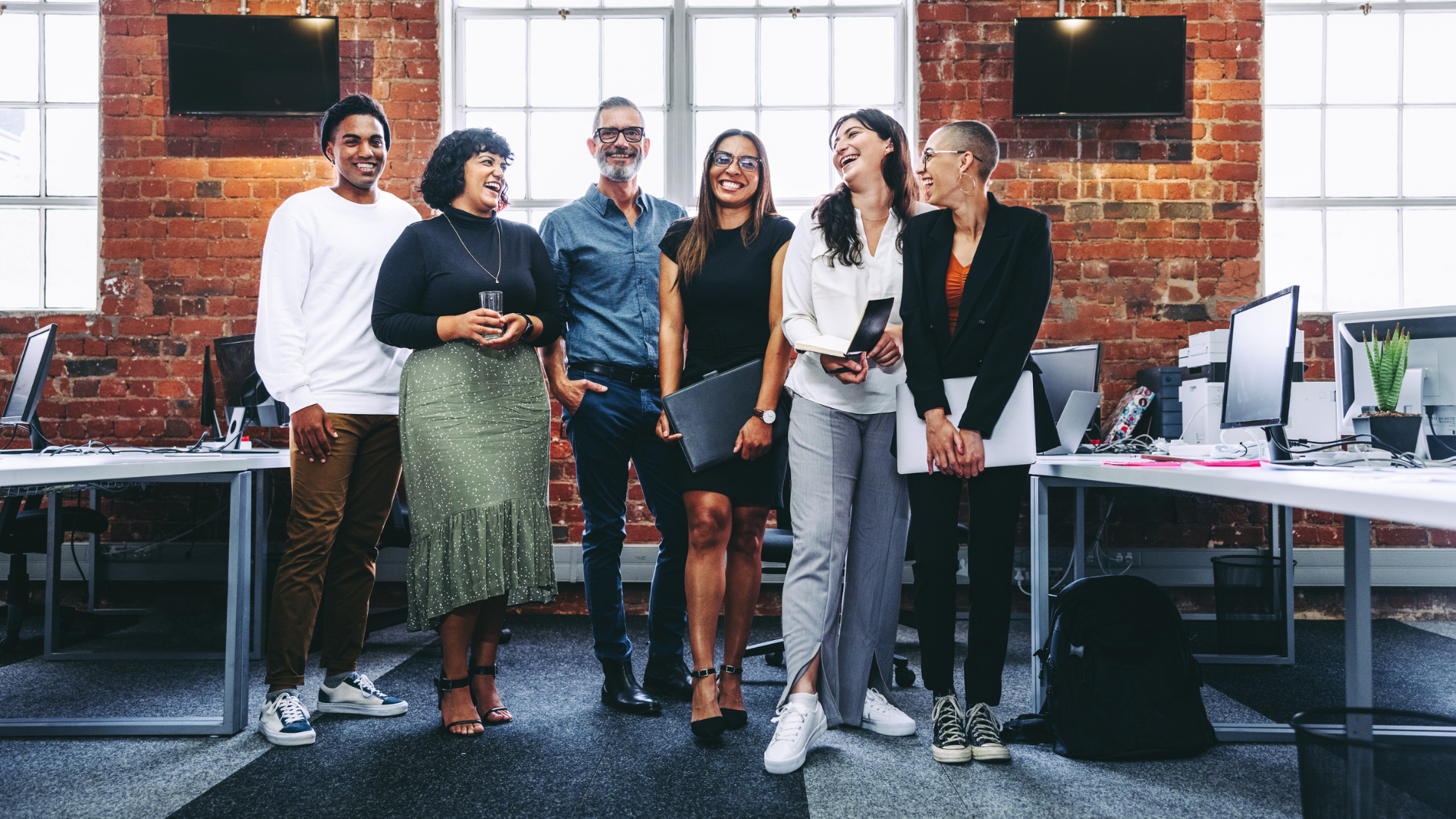 Six diverse business professionals stand together in an open office, smiling and laughing, all dressed in casual clothing.