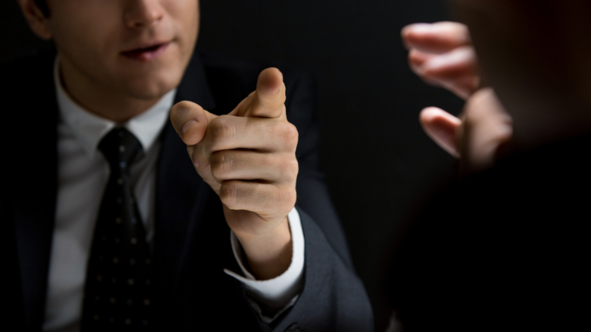 A suited man points assertively at another man, embodying the demeanor of a tyrannical leader in a tense confrontation.