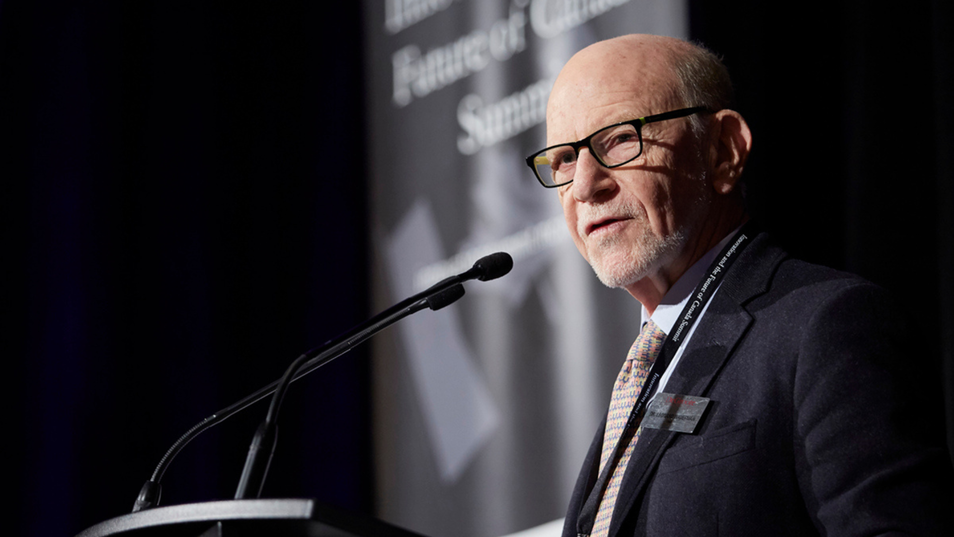 Leonard Waverman, wearing glasses and a suit, speaks confidently at a podium during a formal event.