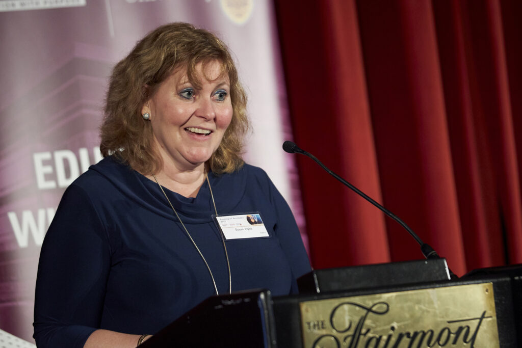 Susan Tighe speaks at a podium, addressing the audience with a red curtain backdrop during the event.