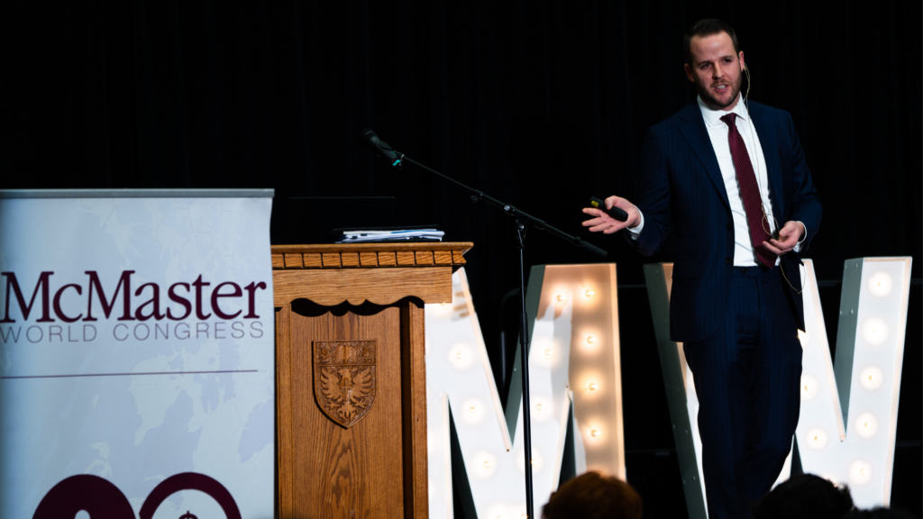 Derek Brenzil, dressed in a suit and tie, presents at a podium, gesturing to his presentation slides that are off screen.