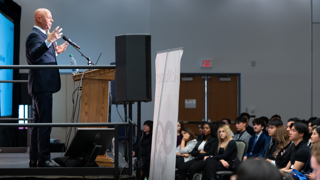 Paul Taylor, in a suit, is seen from the side as he addresses an attentive audience during his speech.