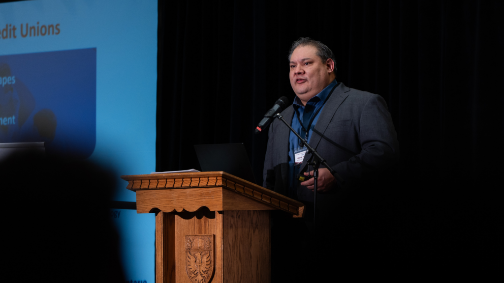 Mark Perkins stands at a podium with a laptop, delivering a presentation.