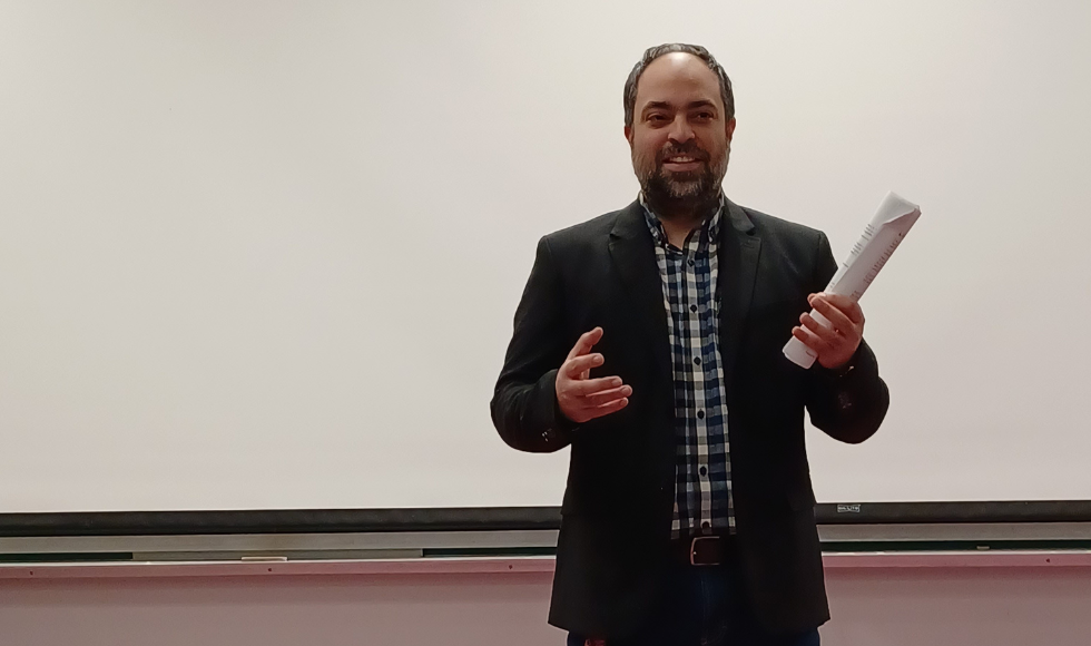 Behrouz Bakhtiari, dressed in a suit, addresses an audience while holding a rolled-up document in his left hand.