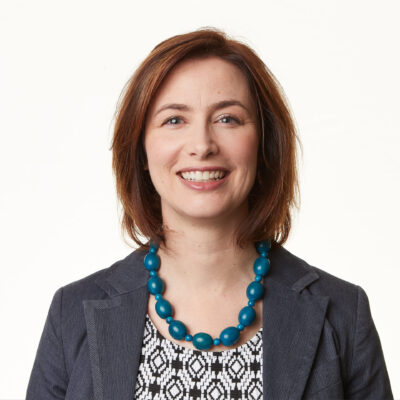 A smiling woman in a business suit, adorned with a blue necklace.