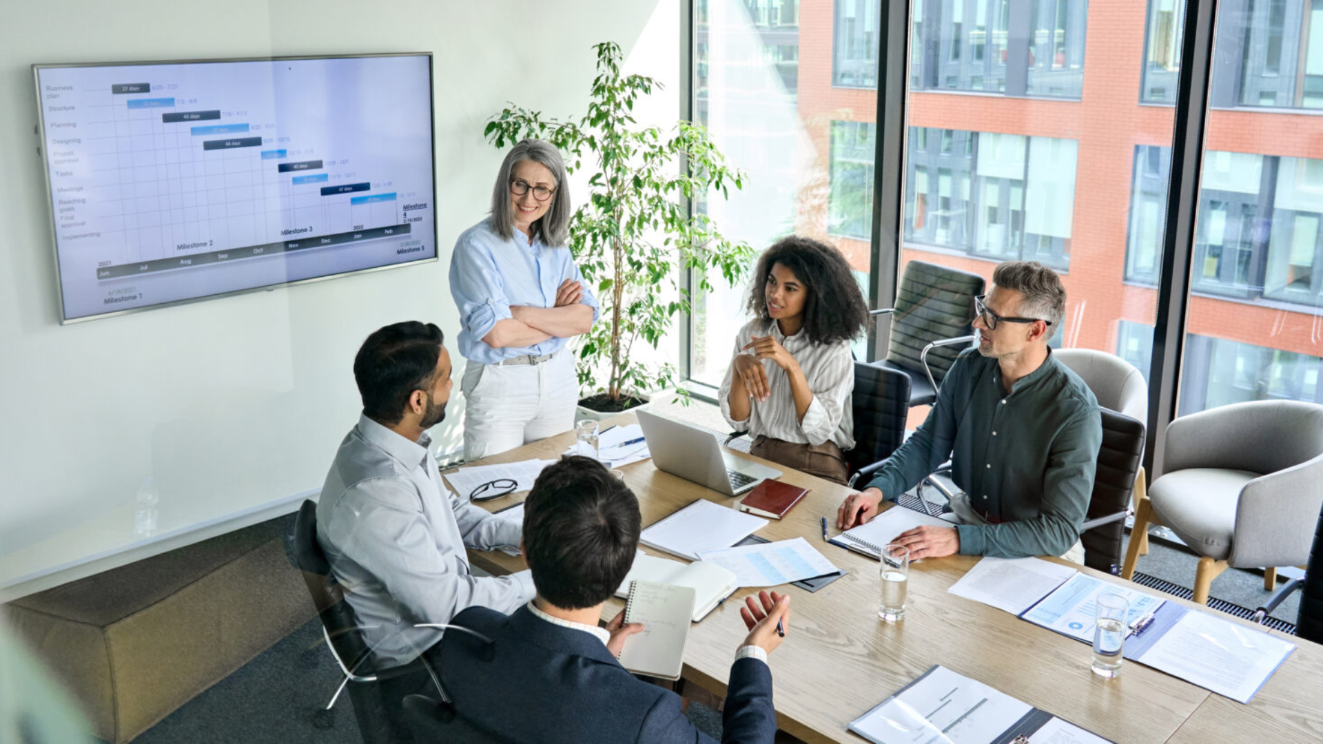 A diverse group of individuals engaged in discussion in a meeting room, with a projector screen displaying information.