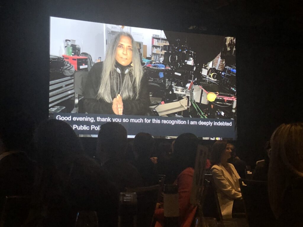 Honouree Deepa Mehta speaks to a crowd, displayed on a large screen at the 34th Annual Testimonial Dinner and Awards.