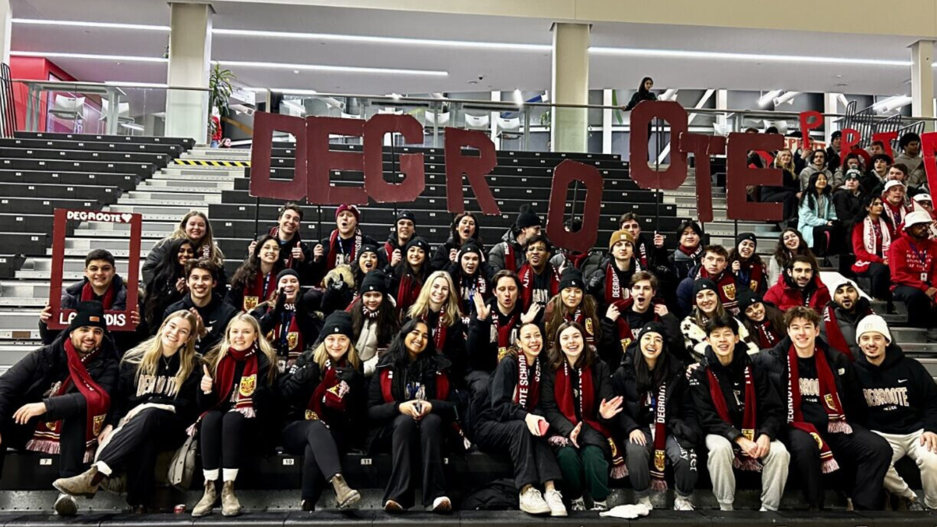 A group of DeGroote JDCC team members on bleachers, proudly displaying a sign that reads 