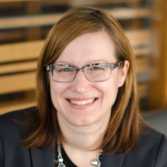 Jennifer Dunk, a woman in glasses and a black blazer, sits down with a warm smile on her face.