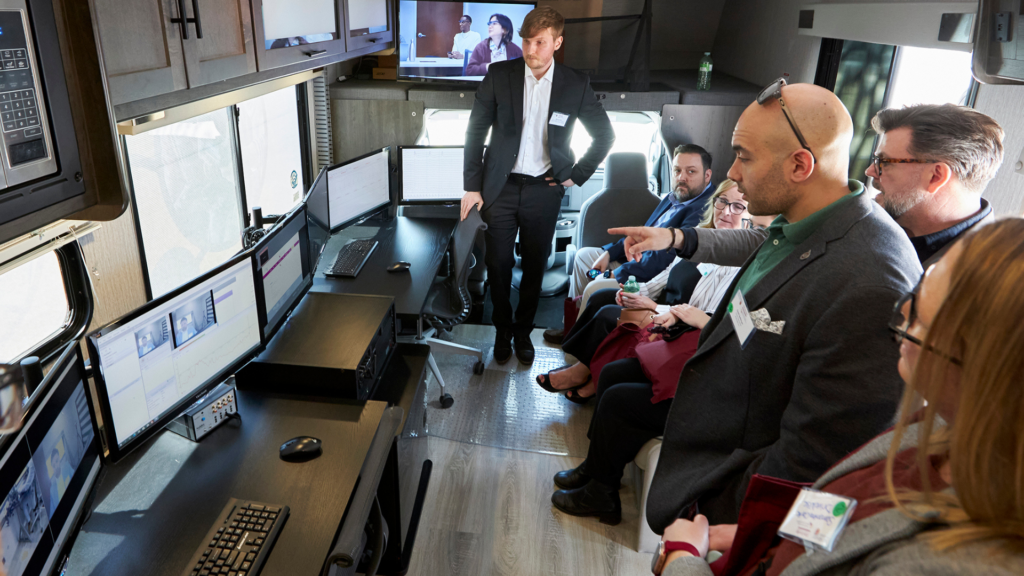 A group of individuals on a bus observes monitors as a research assistant demonstrates neurophysiological tools in a mobile lab.