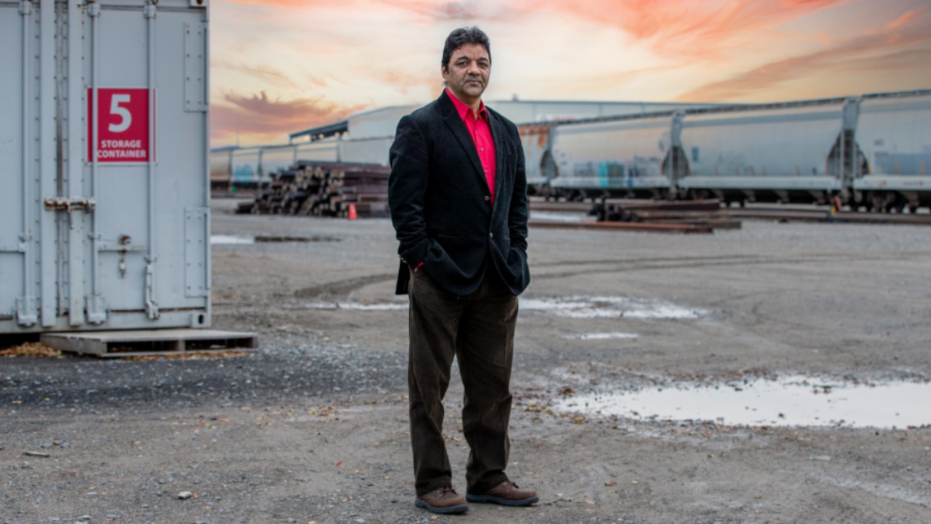 A man in a red shirt and black jacket stands confidently in front of a train at an industrial site, hands in pockets.