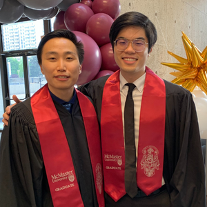 Michael Wong and Victor Yau in graduation gowns and sashes, smiling beside colorful balloons at their graduation celebration.