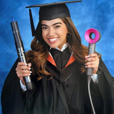 Momina Qureshi smiling in a graduation gown and cap with holding two hairstyling tools.