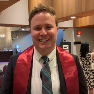 Nicolas Ellis, dressed in a suit and tie, smiles proudly while wearing his graduation sash.