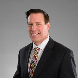 Paul Heinrich, a man in a black suit and tie, smiles warmly for the camera against a neutral background.