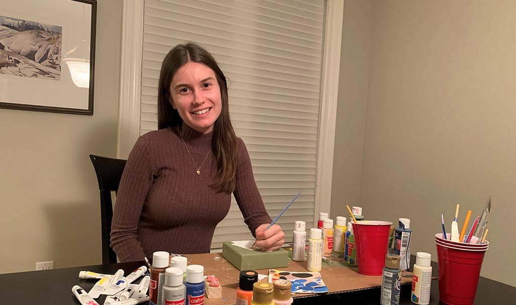 Rachel West smiles at a table filled with art supplies, holding a paintbrush beside a colorful box of paints.