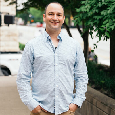 Tal Dehtiar, dressed in a blue shirt and tan pants, smiles while standing on a sidewalk.