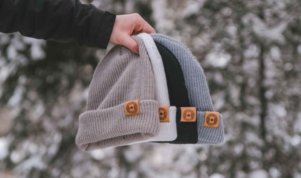 A person holds four colored toques—beige, white, black, and grey—on an outstretched arm.