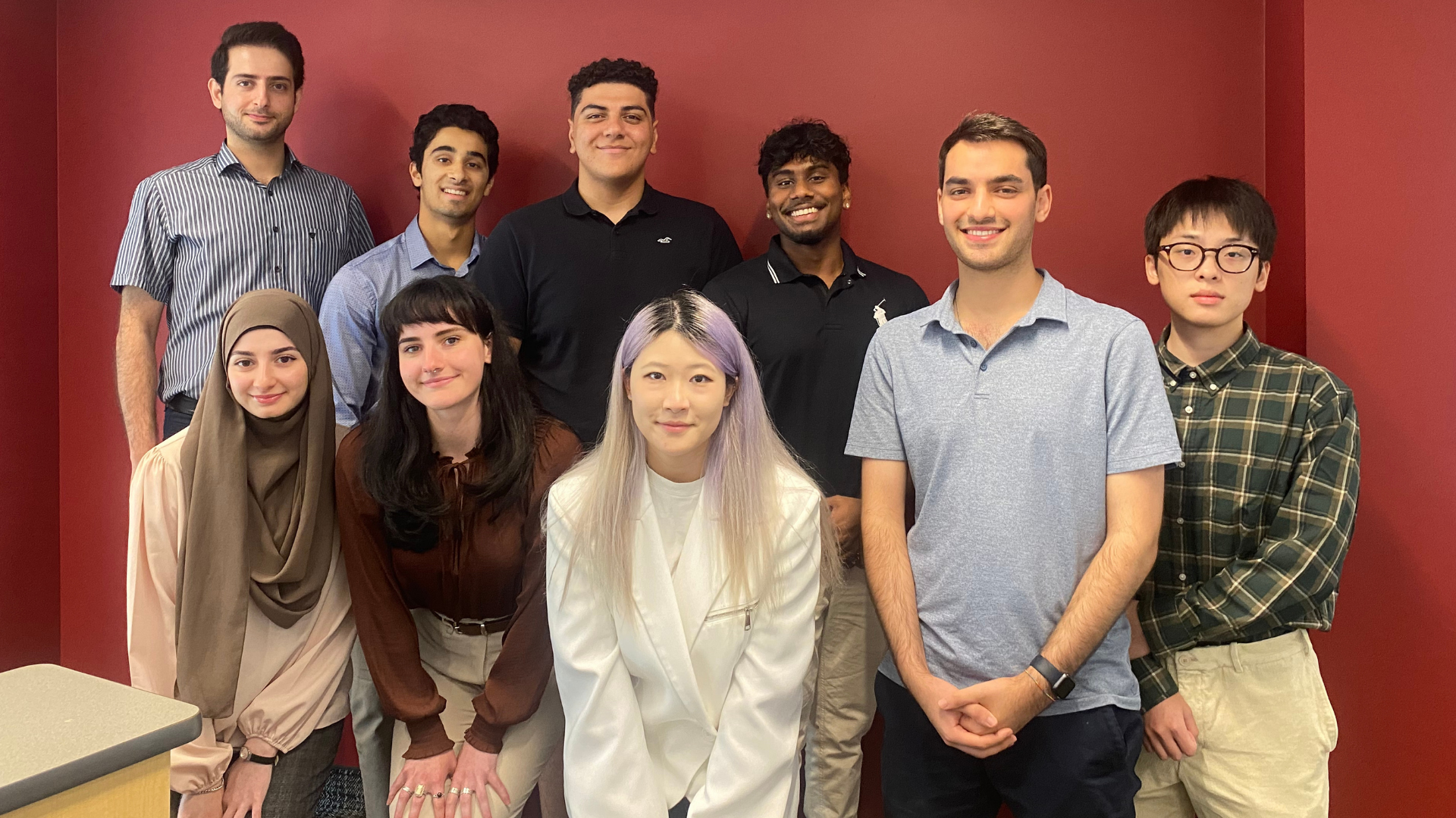 A cheerful group of DeGroote student researchers stands in front of a bold red wall.