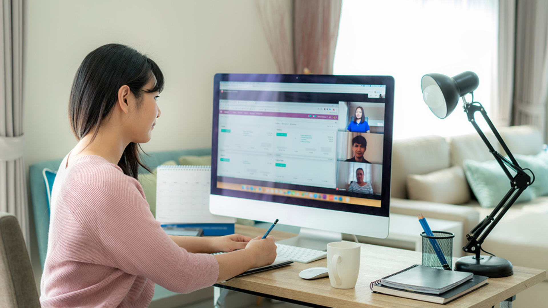 A woman seated at a desk, engaged in a video conference while taking notes on her computer.