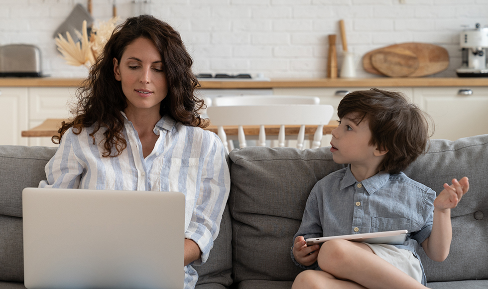 A woman engages with her laptop on the couch, as a small child beside her attempts to capture her focus.