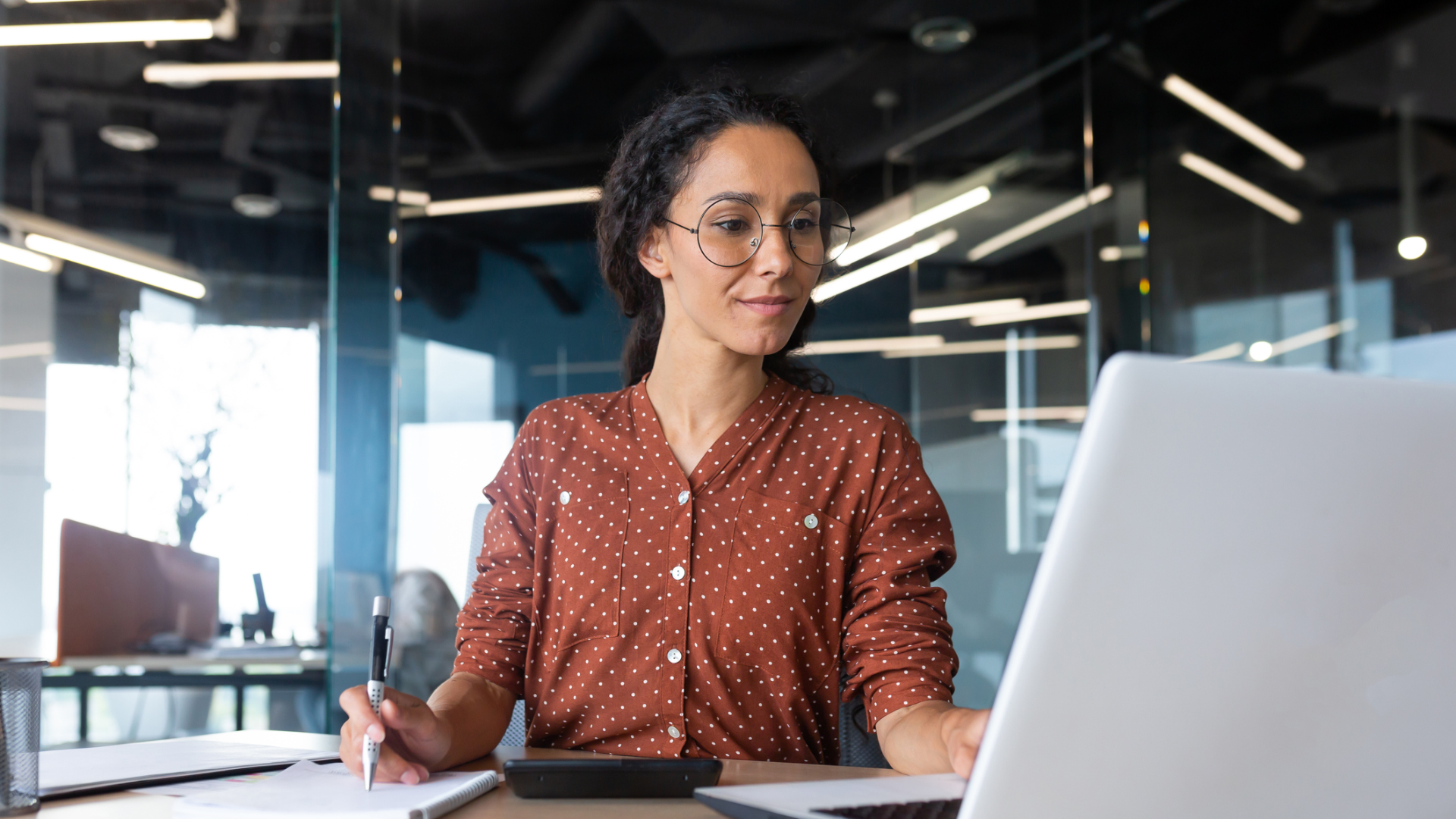 A woman wearing glasses is focused on her laptop, engaged in work at a desk in a well-lit environment.