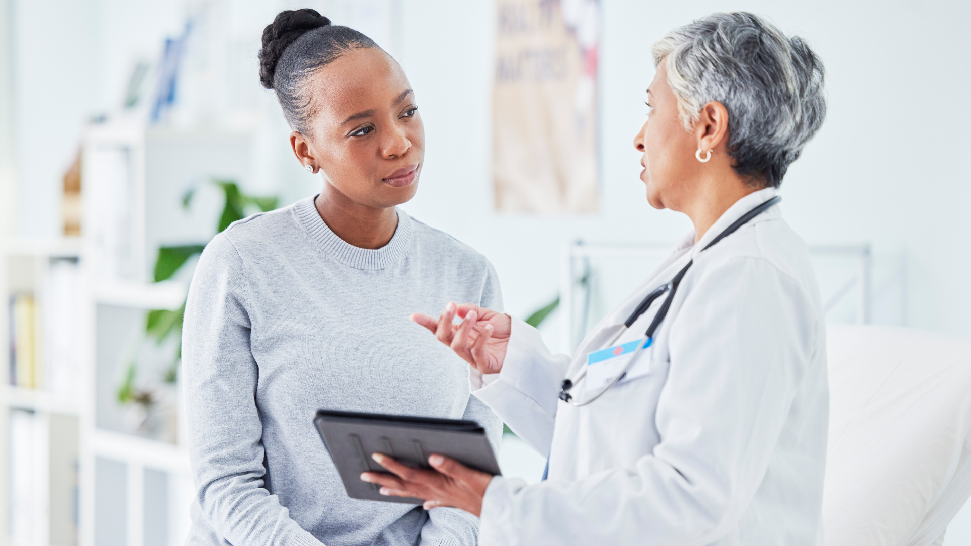 A woman consults with a doctor while holding a tablet, engaged in a discussion about her health.