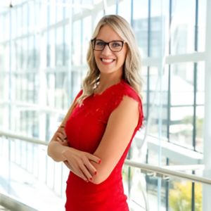 Jessica Bernat, wearing glasses and a red dress, smiles while standing in front of a window.