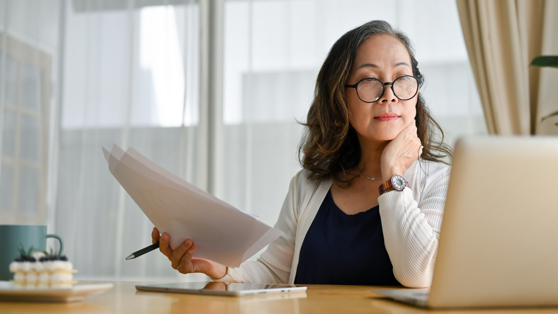 A senior woman wearing glasses sits at a table, diligently working on her laptop.